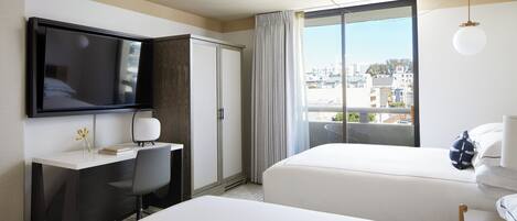 A hotel room with a TV, desk, chair, two beds, a window with curtains, and a view of buildings.