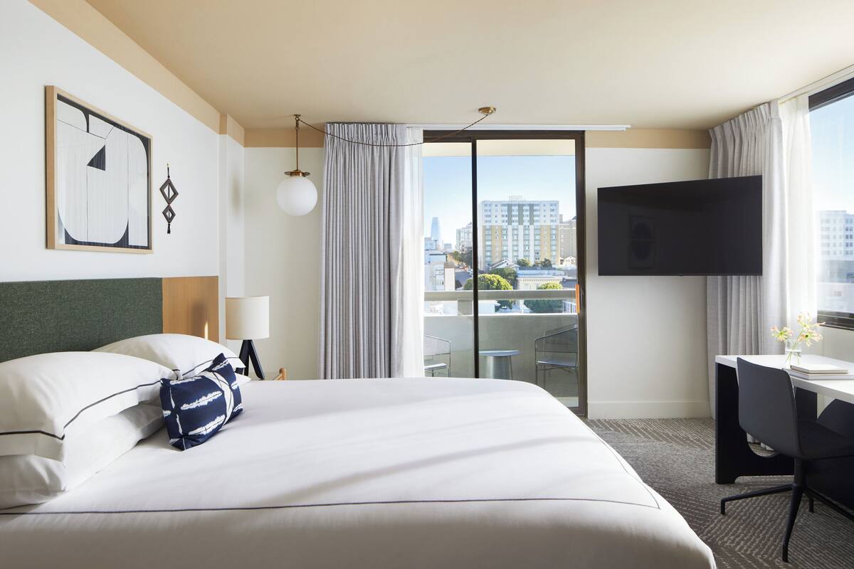 A hotel room with a bed, a desk, a TV, and a view of the cityscape.