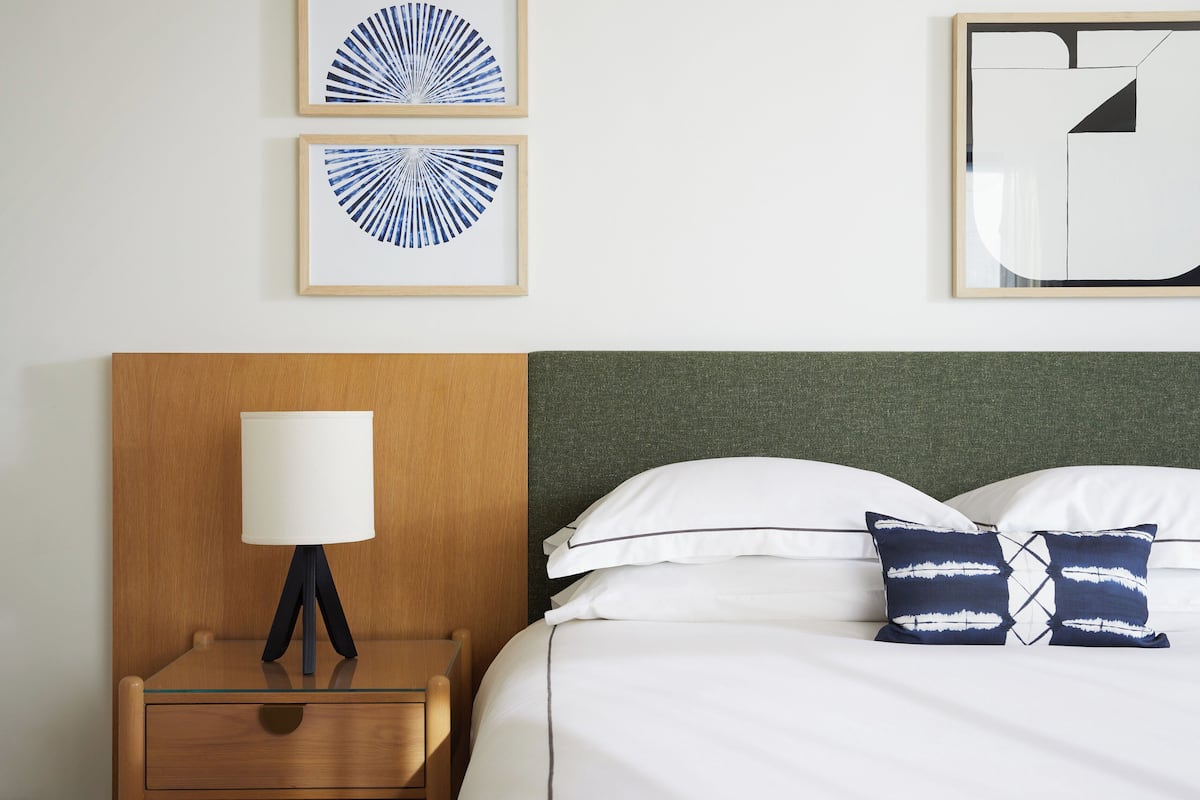 A neatly arranged hotel room with a wooden nightstand, a bedside lamp, a bed with white bedding and blue patterned pillows, and two framed abstract wall art pieces.