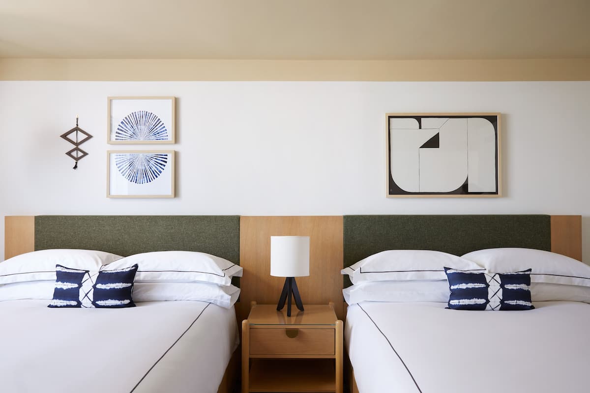 A hotel room with two beds, a nightstand with a lamp, and two framed artworks on the wall.