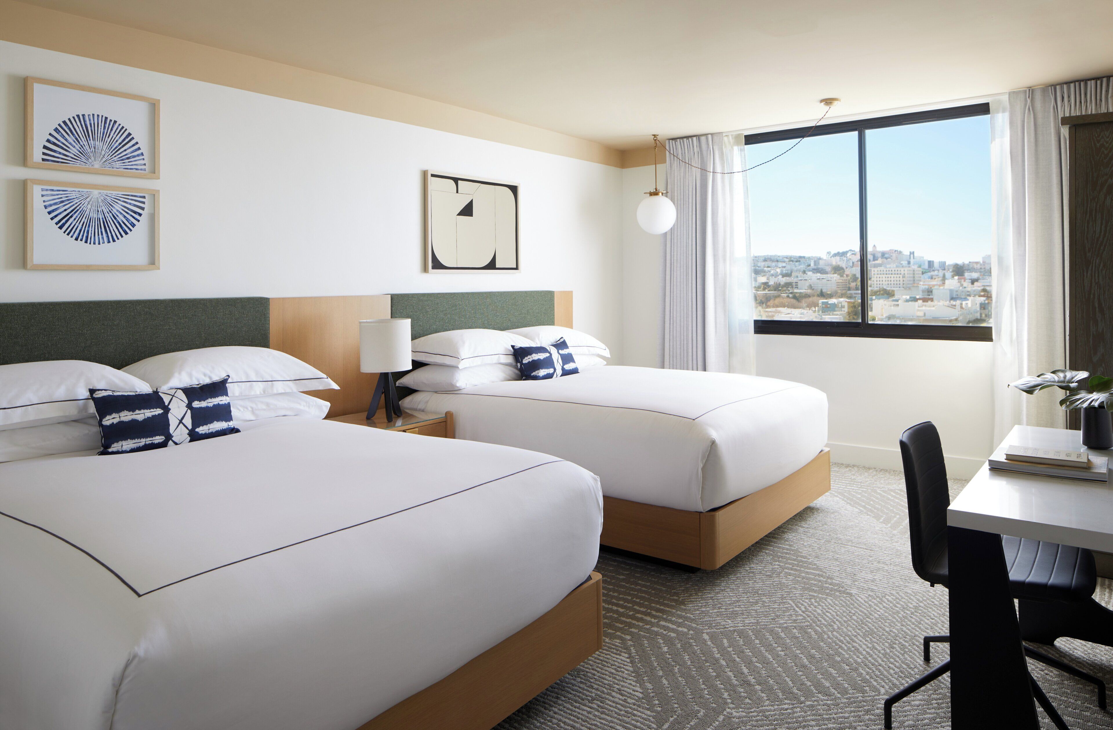 A hotel room with two beds, a desk, a chair, a lamp, and a view of the city.
