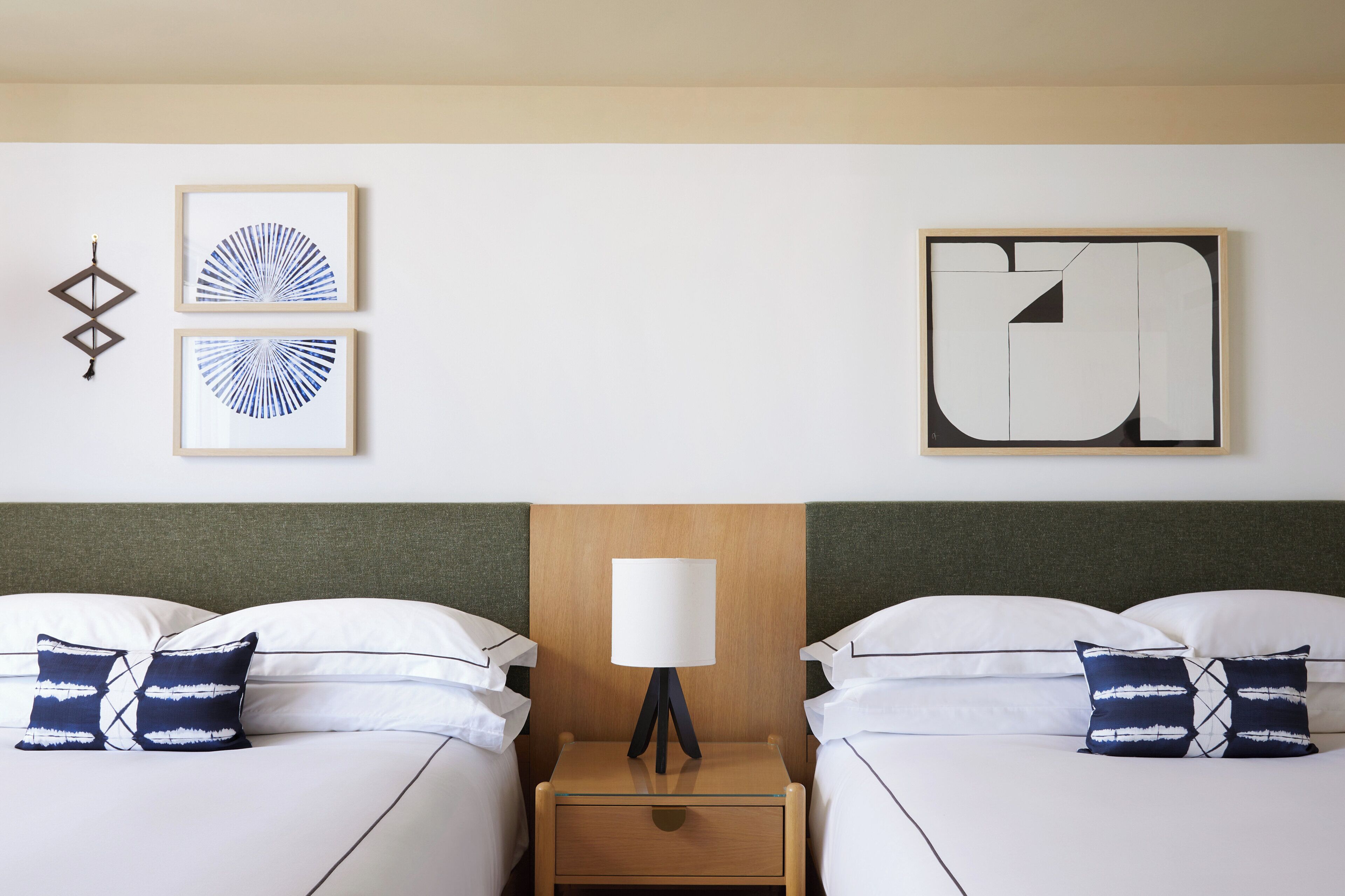 Ein Hotelzimmer mit zwei Betten, einem Nachttisch mit Lampe und zwei gerahmten Kunstwerken an der Wand.