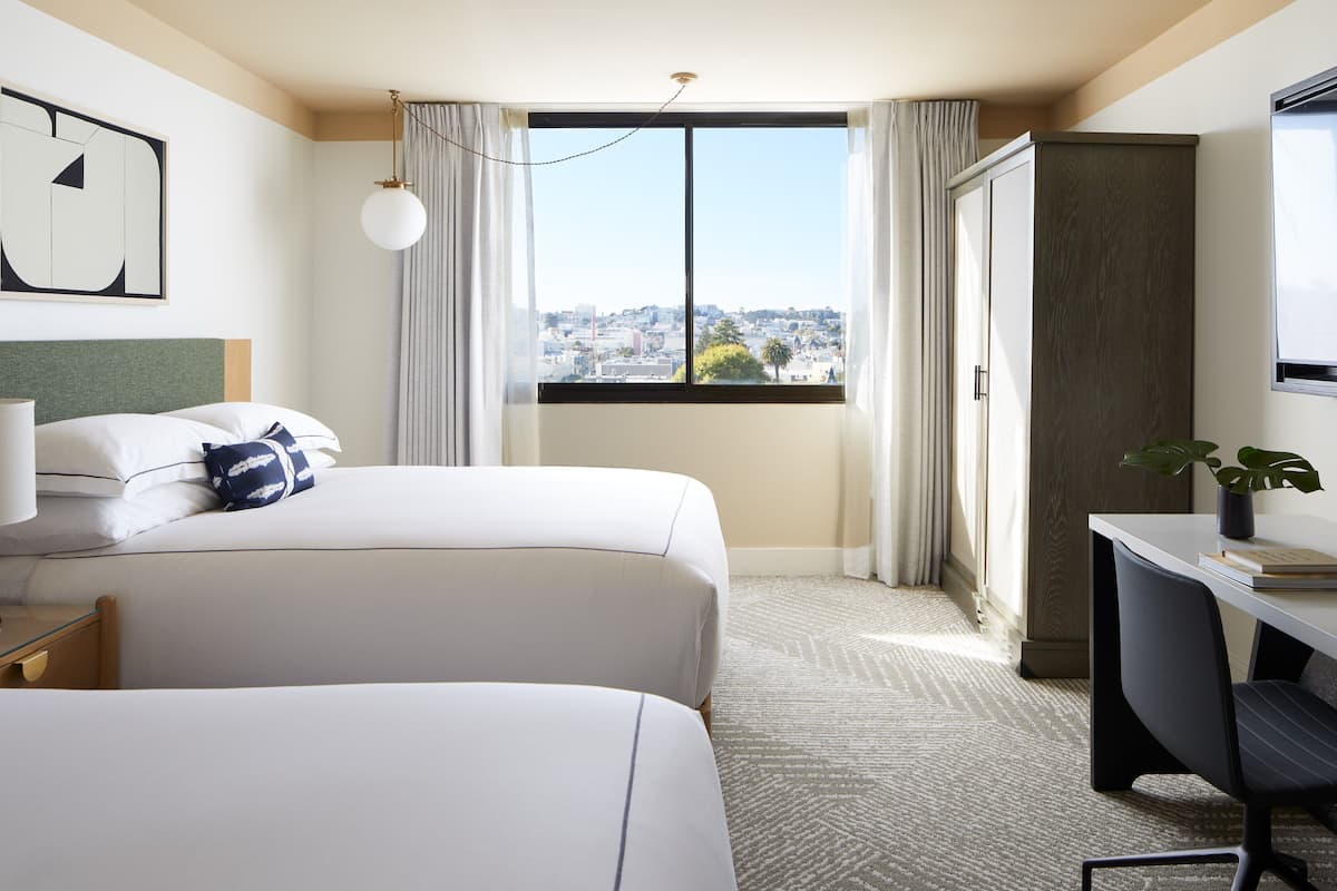 A hotel room with two beds, a desk, a chair, a large window with a city view, and a wardrobe.