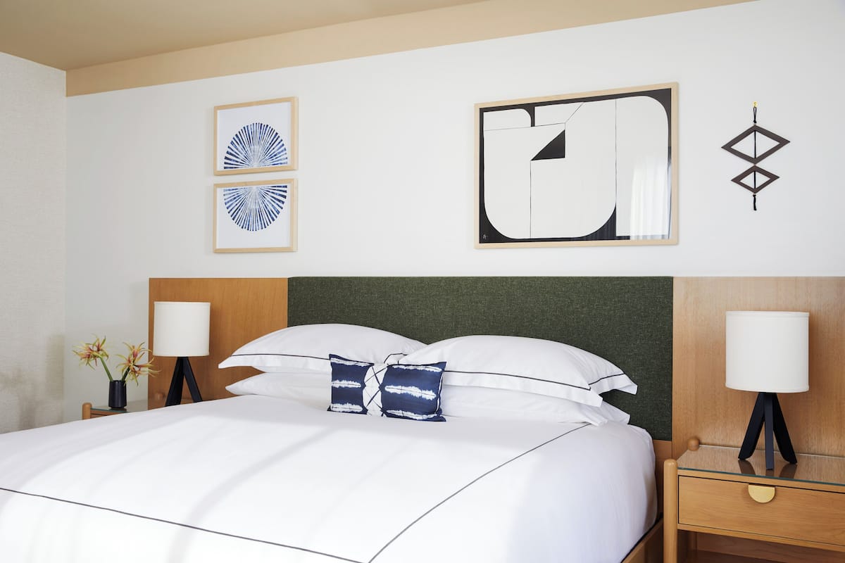 A neatly made bed with white linens and two decorative pillows, a bedside table with a lamp, and wall art including framed abstract designs.