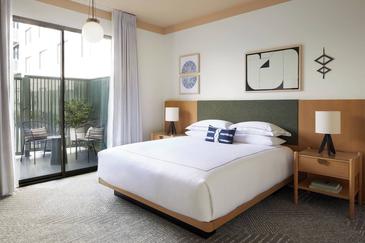 A hotel room with a large bed, two bedside tables with lamps, a view of the outside balcony, and artwork on the walls.