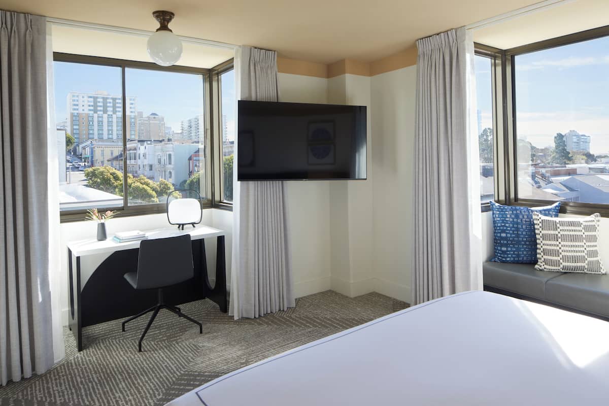 A hotel room with a large window, a desk, a chair, a bed, and a flat-screen TV.