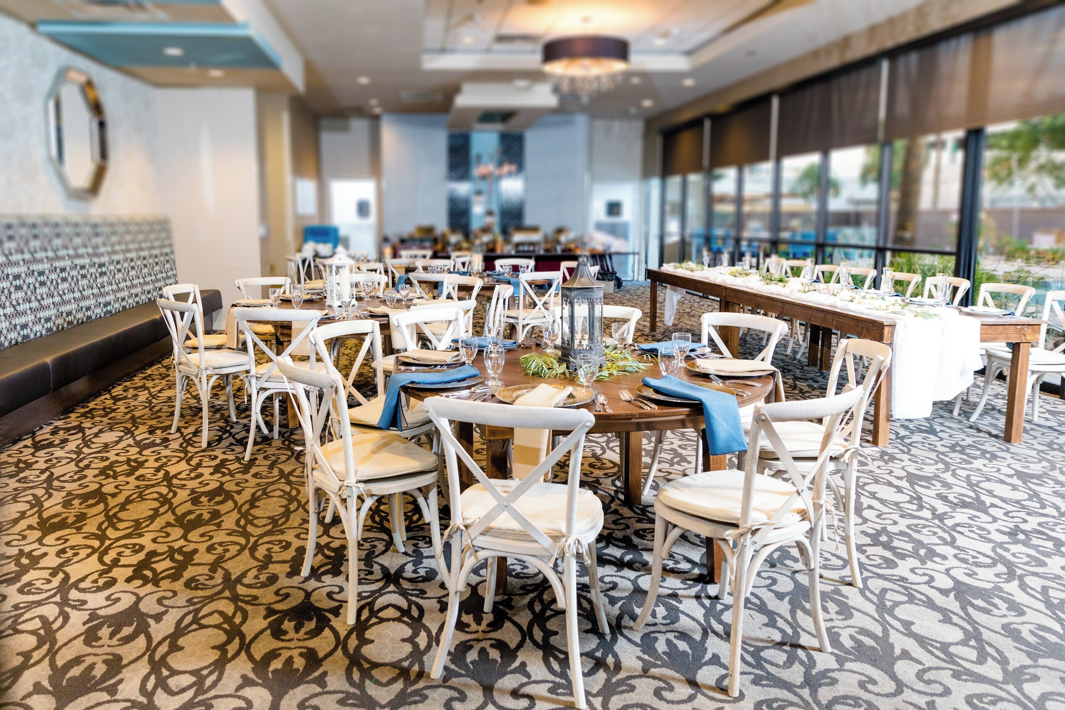 A hotel dining area with multiple tables and chairs, large windows, and a patterned carpet.