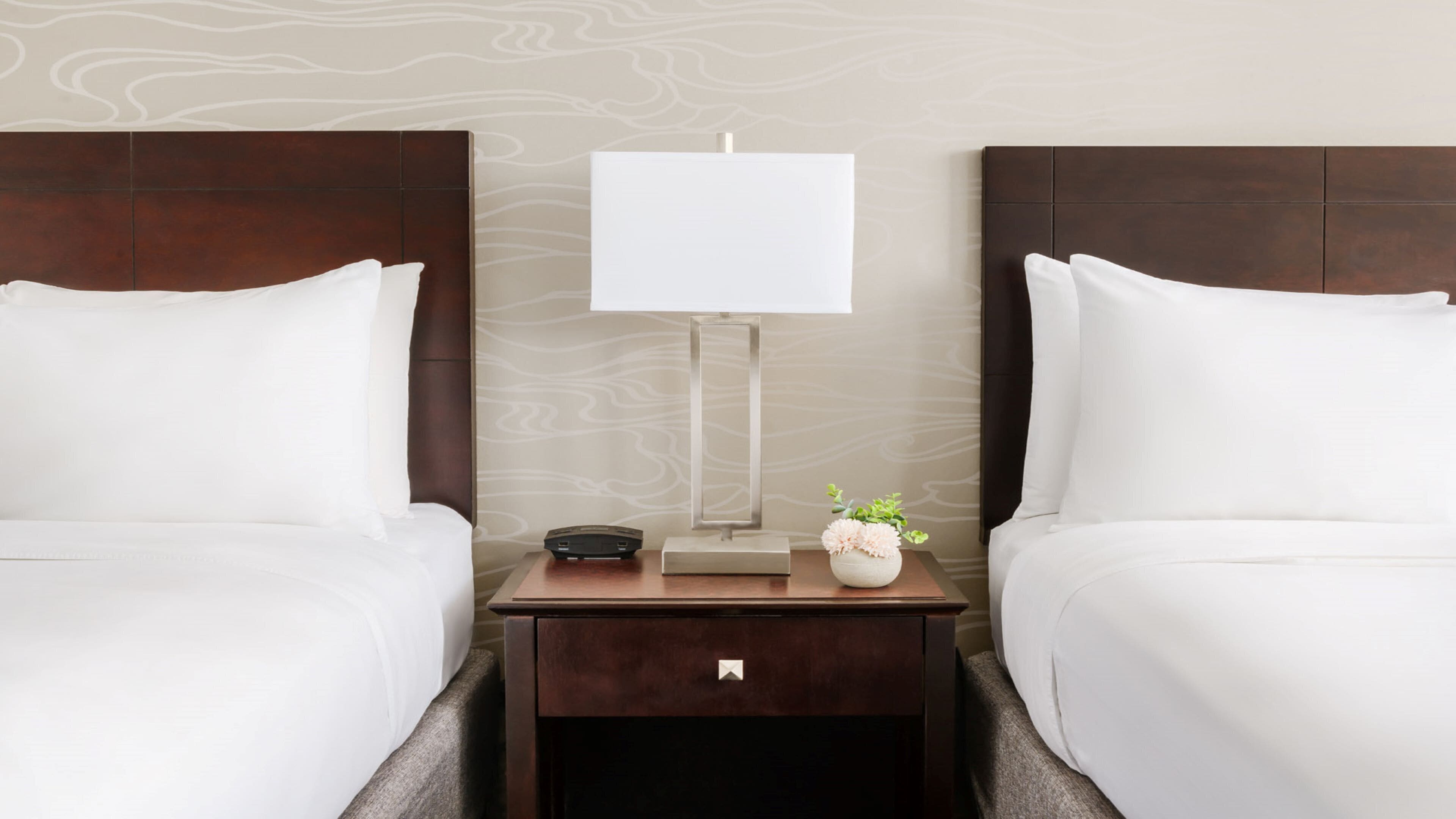 A hotel room with two beds, a nightstand with a lamp, a phone, and a small vase with flowers.