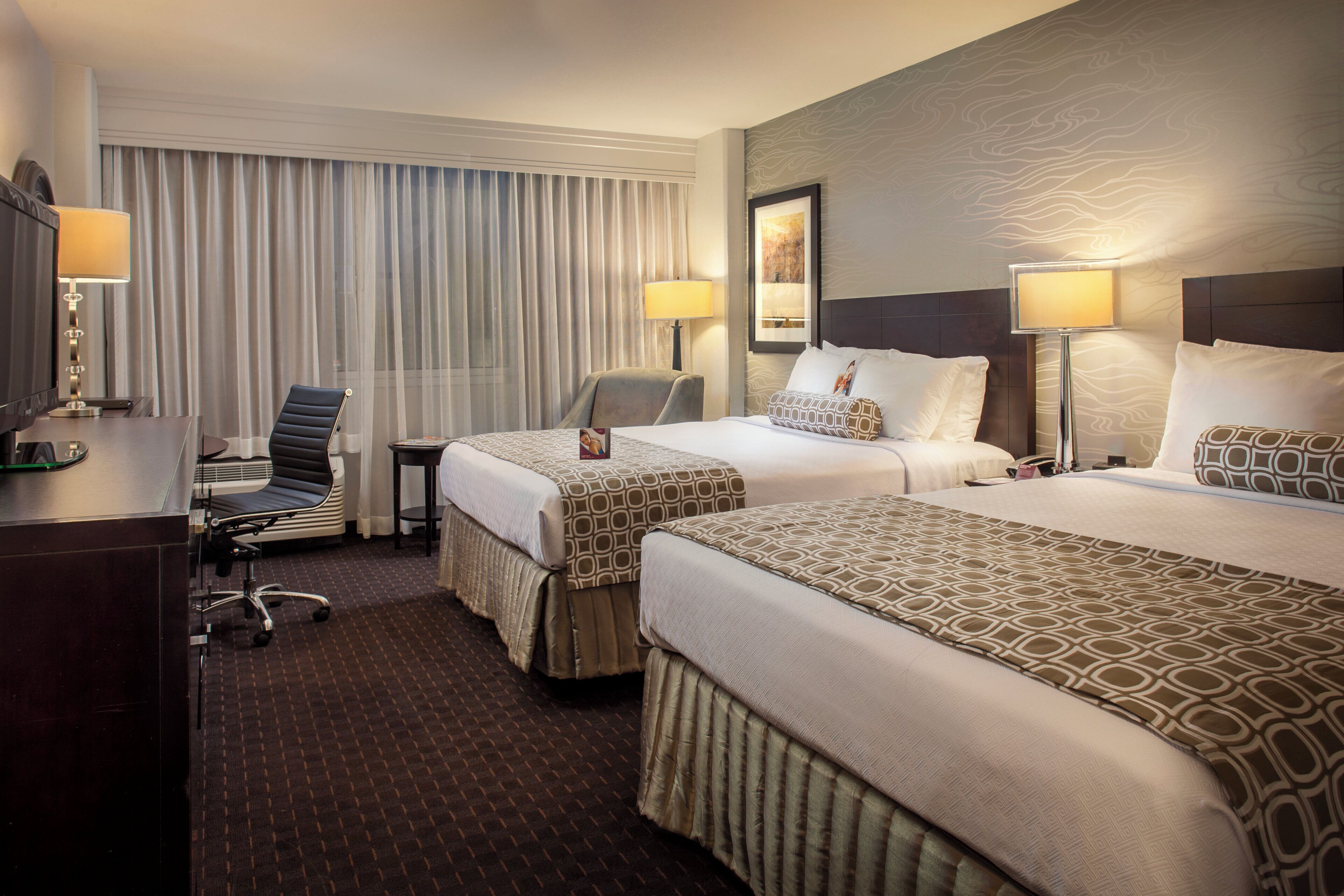 A hotel room with two beds, a desk, a chair, and a window with curtains.
