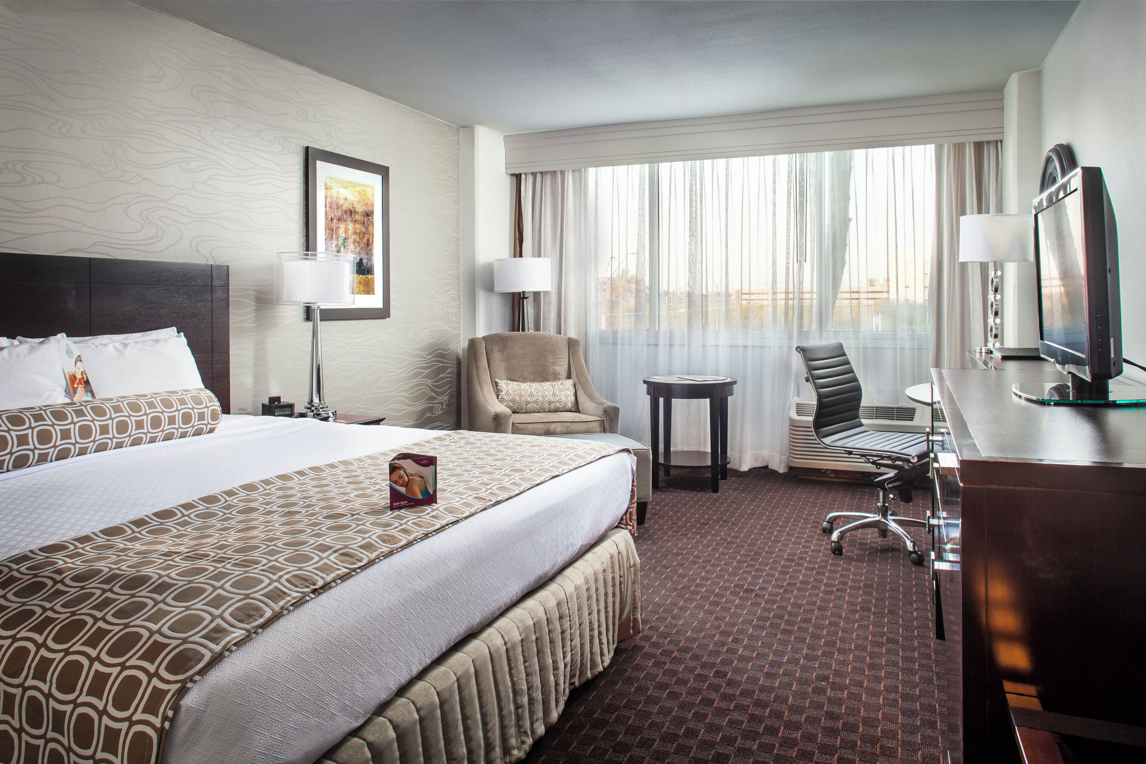A hotel room with a large bed, a desk with a computer, a chair, a TV, and a window with curtains.