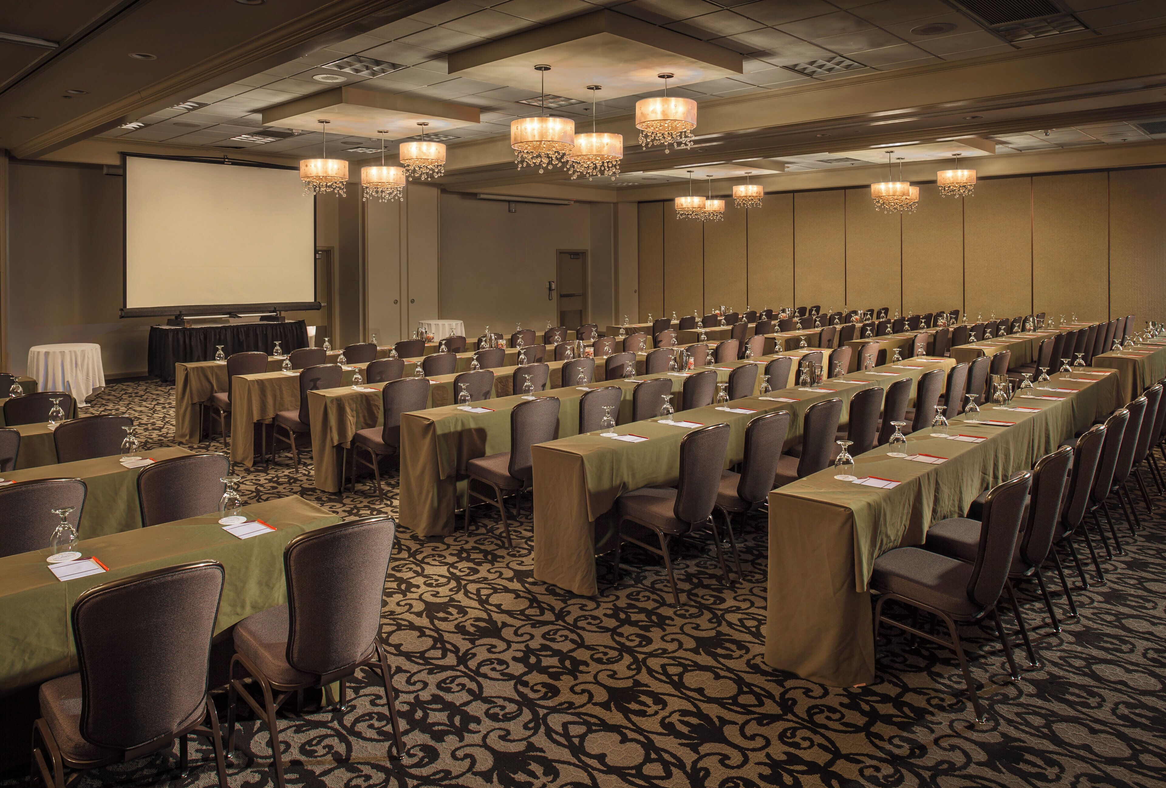 A conference room with rows of tables and chairs, a projector screen, and chandeliers.