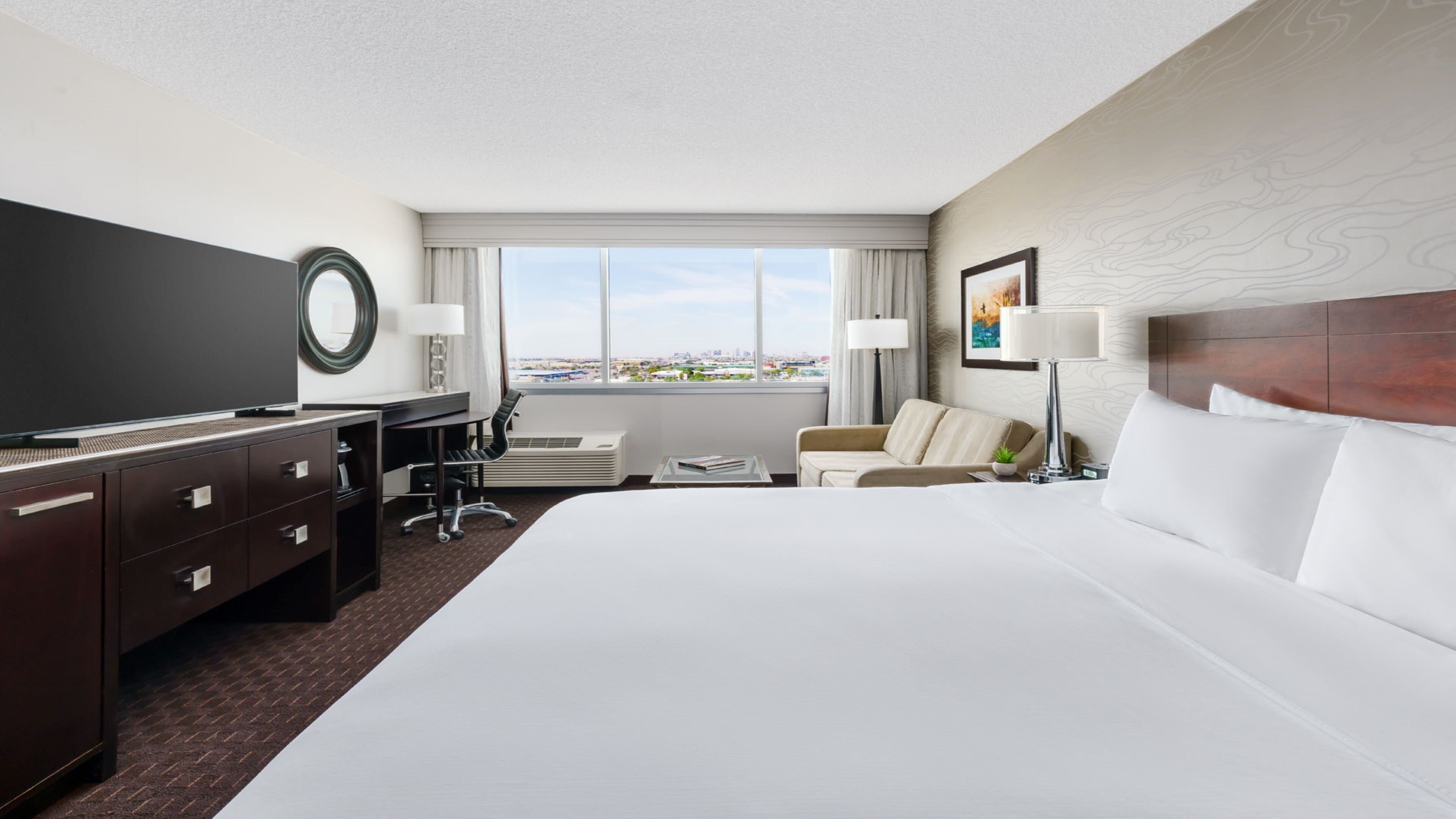 A hotel room with a large bed, a desk with a flat-screen TV, a chair, a sofa, and a window with a city view.