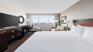 A hotel room with a large bed, a desk with a flat-screen TV, a chair, a sofa, and a window with a city view.