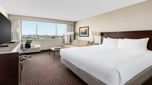 Habitación de hotel con una cama grande, un escritorio con silla, un sofá, un televisor y una ventana con vistas a la ciudad.