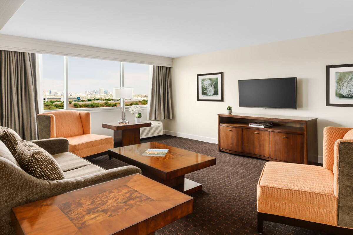Habitación de hotel con sofá, sillón, mesa de centro, televisor y una obra de arte en la pared.