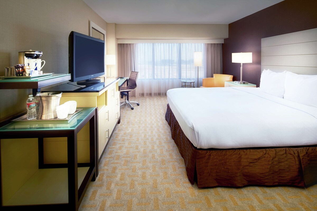 Habitación de hotel con cama, escritorio, computadora, silla, lámpara y ventana con cortinas.