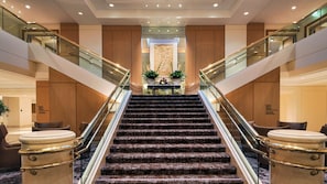 Una imponente escalera de mármol conduce a una zona de recepción con un muro decorativo y plantas en macetas.