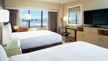 A hotel room with two beds, a desk with a flat-screen TV, a chair, and a view of the airport.
