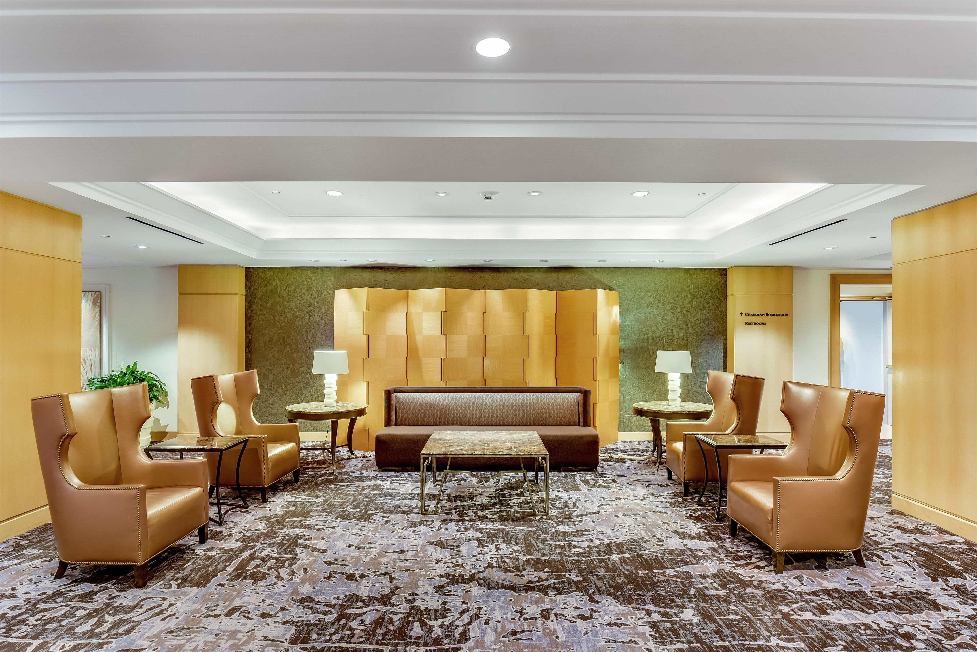A hotel lobby with a central seating area, a large sofa, and multiple armchairs. There are tables with lamps, a plant, and a wall with directional signs.