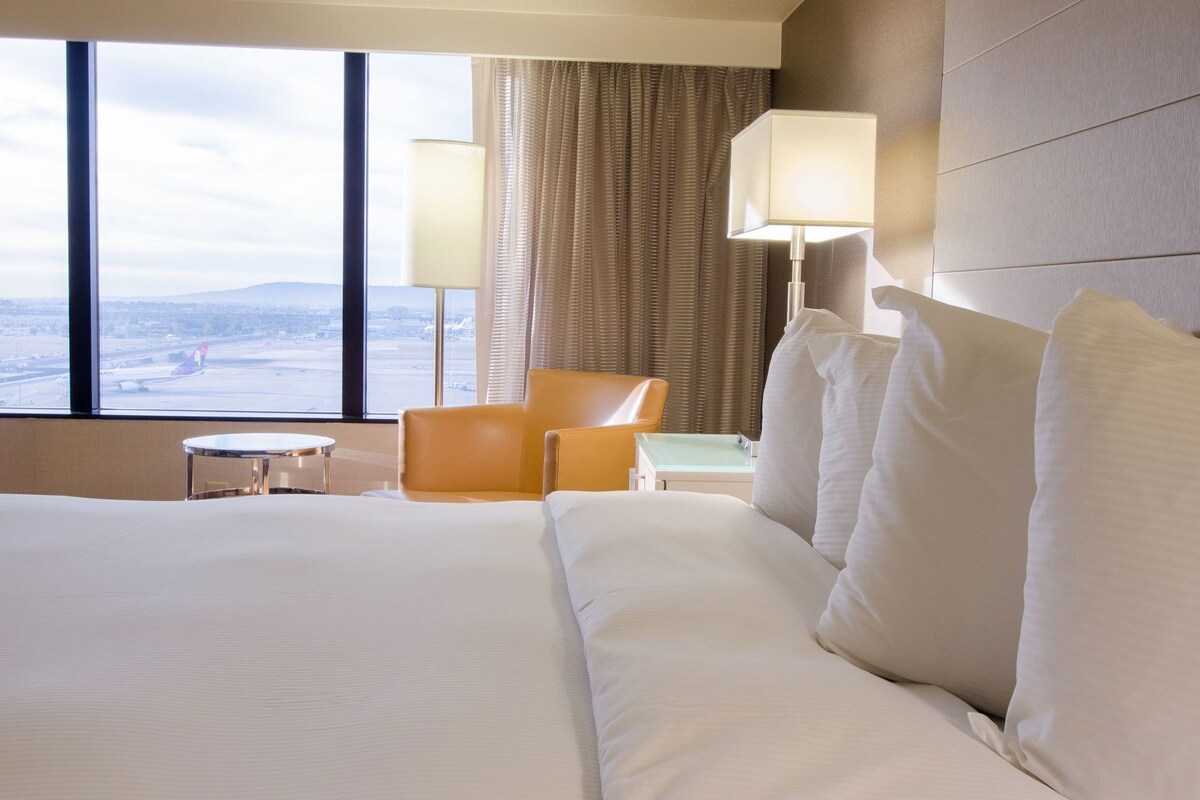 Een hotelkamer met een groot bed, twee stoelen, een klein tafeltje en uitzicht op een landschap door het raam.
