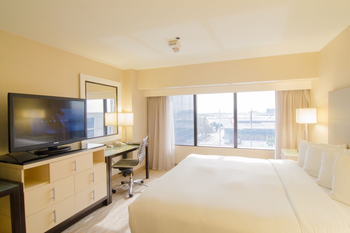 Habitación de hotel con una cama grande, un escritorio con silla, televisión y vistas a la ciudad a través de amplios ventanales.