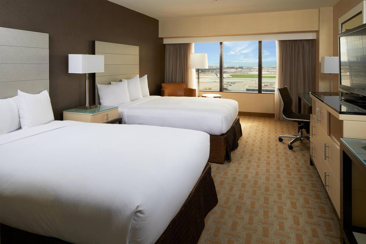 A hotel room with two beds, a desk, a chair, a TV, and a view of the airport.