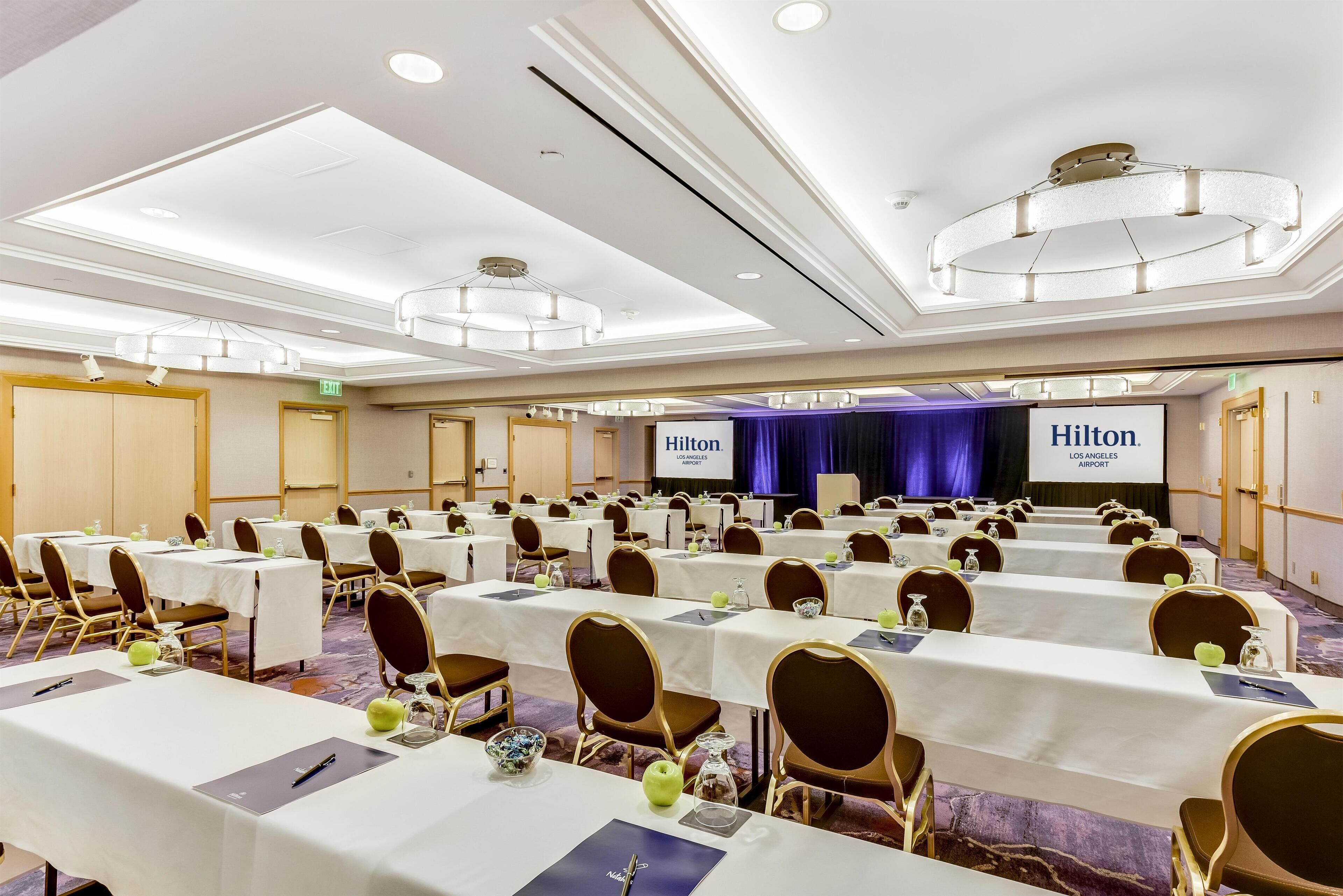 A hotel conference room with multiple tables, chairs, and a stage with a screen.