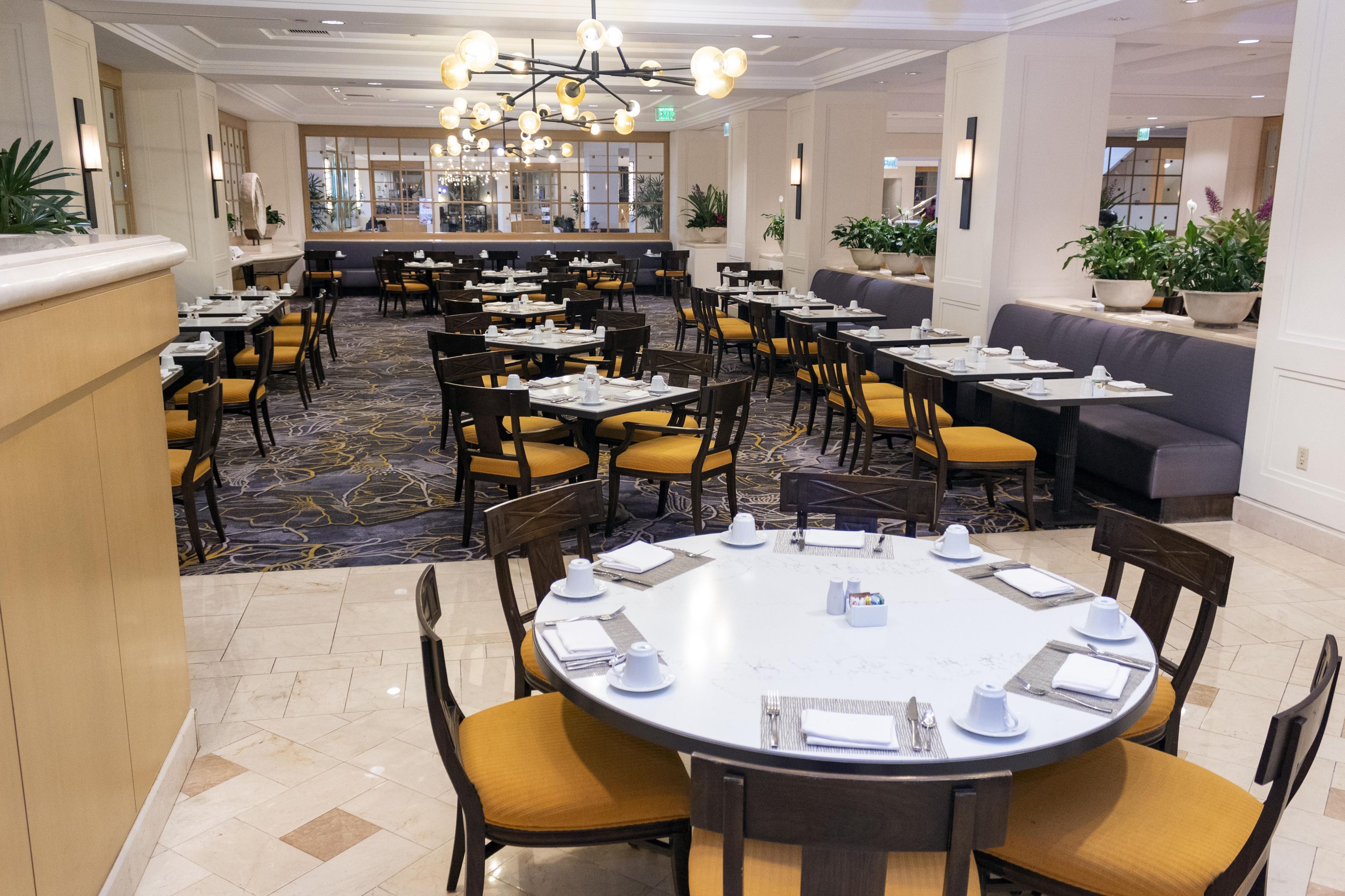 A hotel dining area with multiple tables and chairs, a central seating area with sofas, and a patterned carpet.