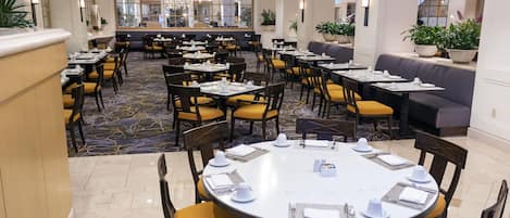 A hotel dining area with multiple tables and chairs, a central seating area with sofas, and a patterned carpet.