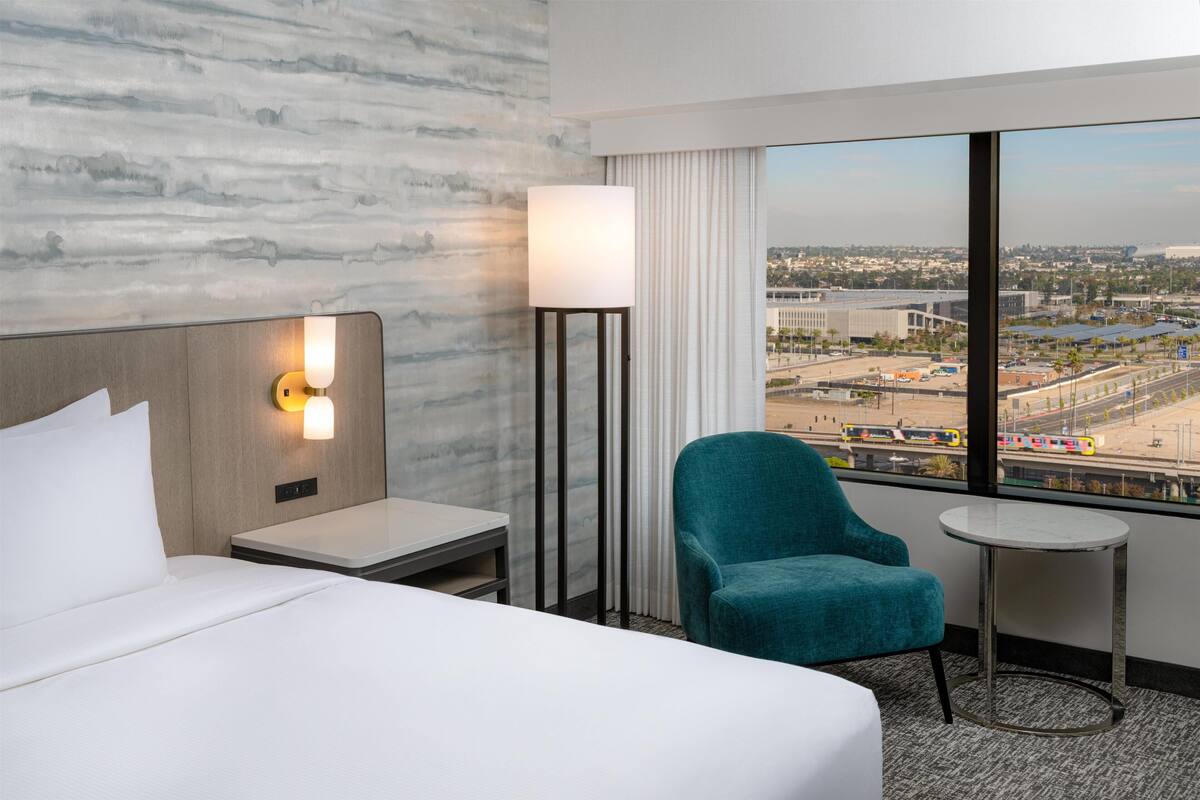 A hotel room with a bed, a nightstand, a lamp, a chair, a small table, and a large window with a city view.