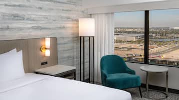 A hotel room with a bed, a nightstand, a lamp, a chair, a small table, and a large window with a city view.