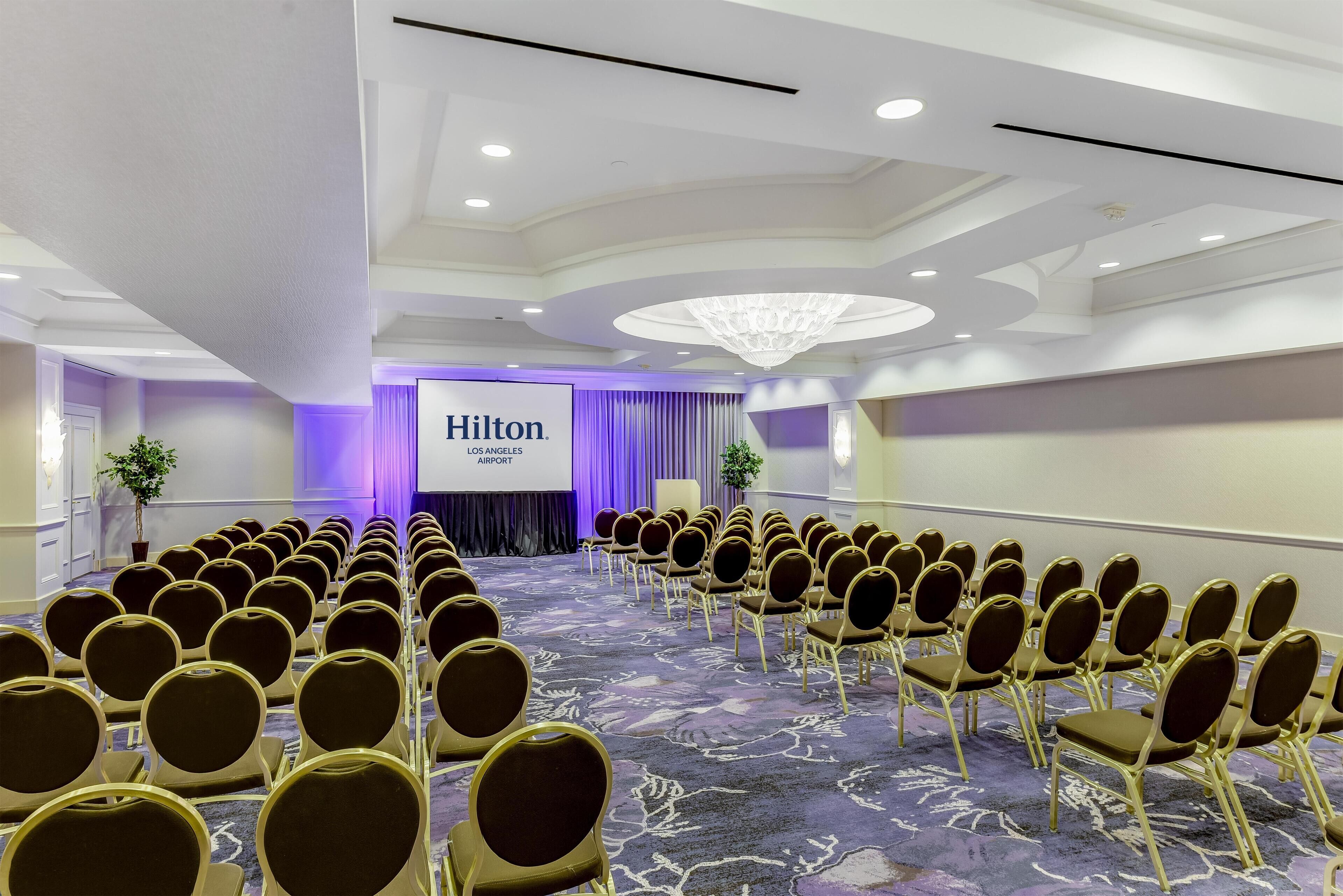 Ein Konferenzraum mit Reihen von Stühlen, einer Projektionsleinwand mit der Aufschrift 'Hilton Los Angeles Airport' und einer Kristallleuchtre.