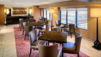 A hotel lobby with a reception desk, multiple seating areas with tables and chairs, and large windows offering a view of the cityscape.