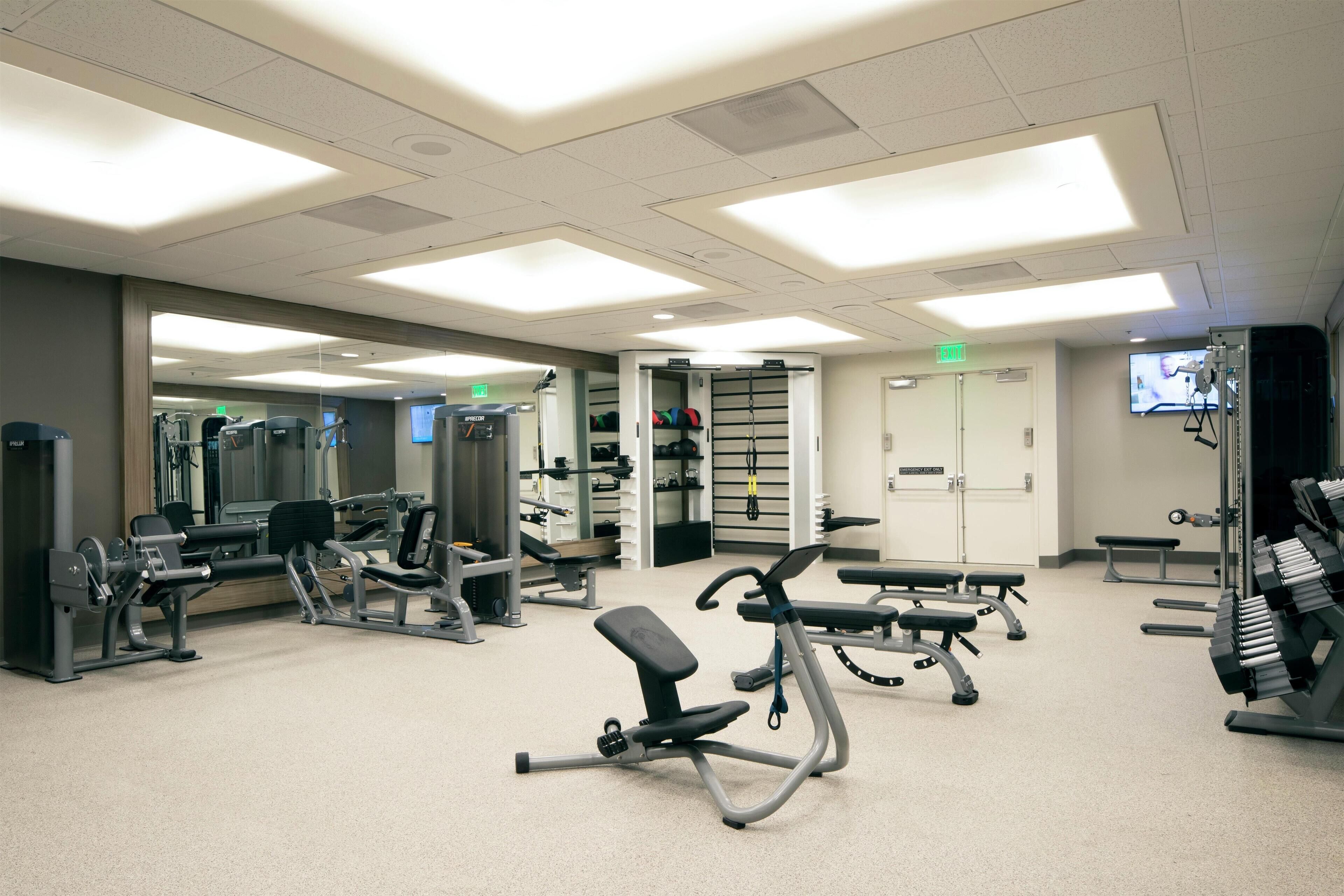 Een sportschool met diverse fitnessapparaten, banken en een spiegelwand.