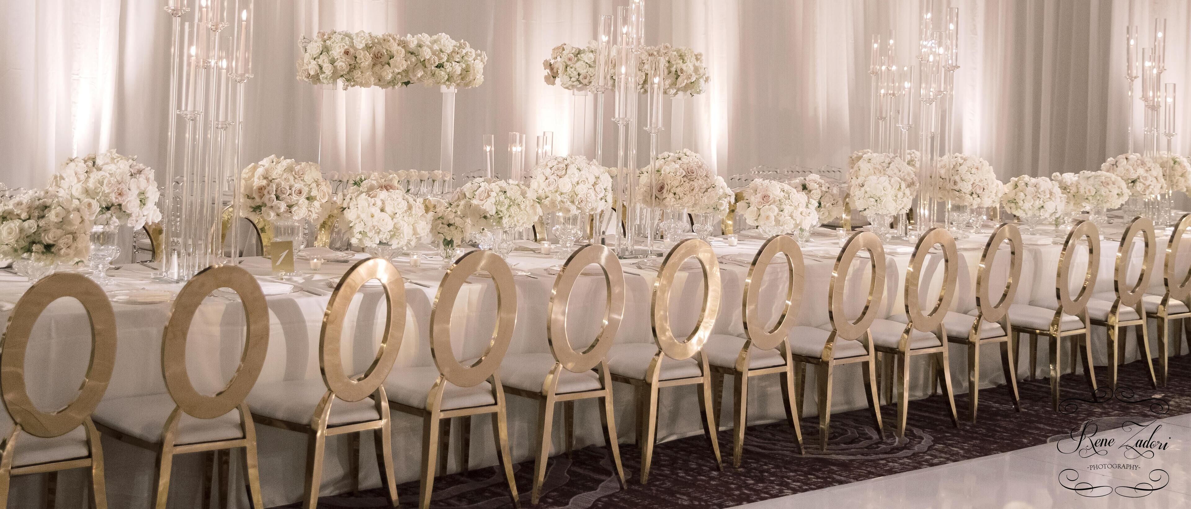 Een lange feesttafel met gouden stoelen, witte tafelkleden en bloemrijke decoraties.