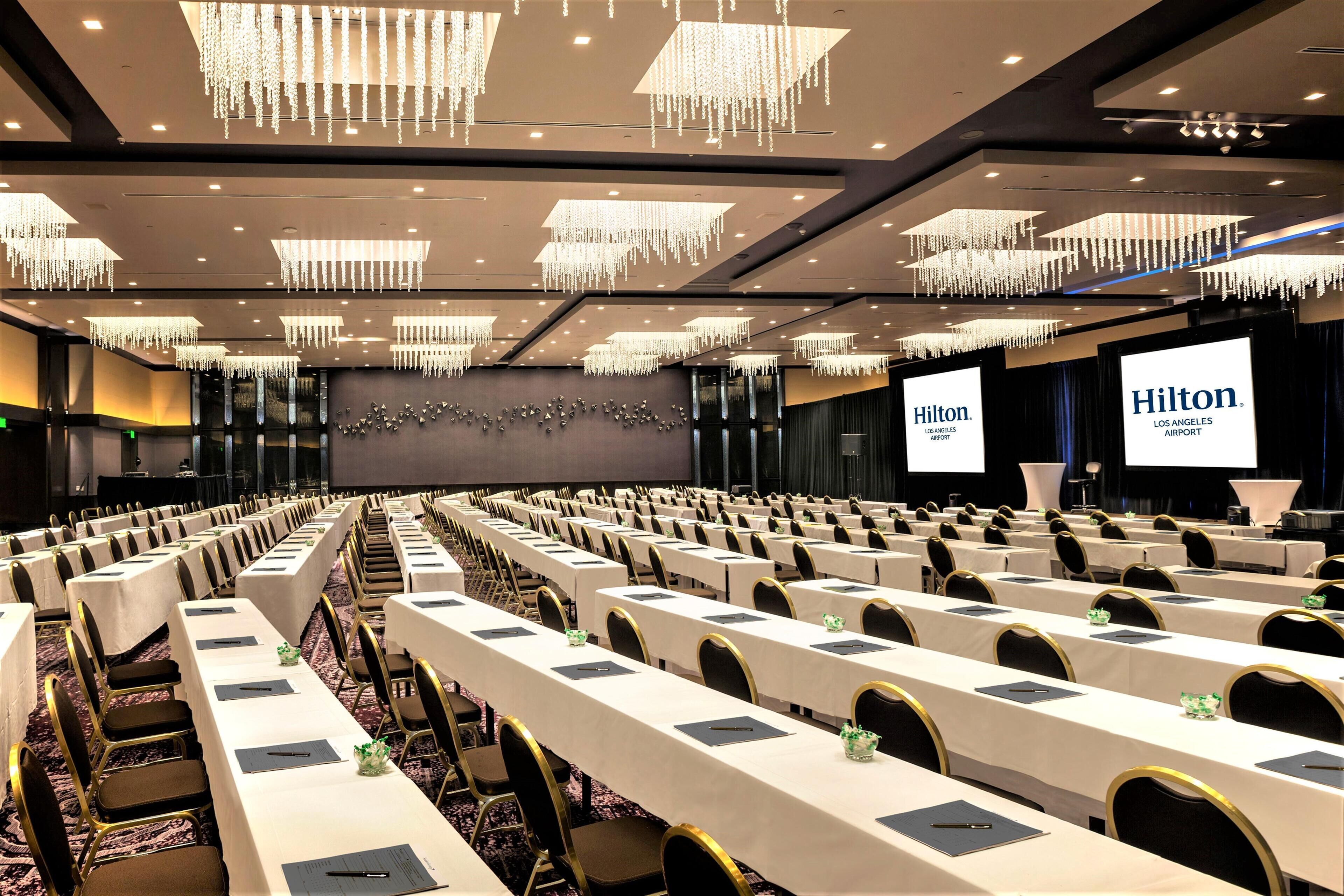 Une salle de conférence avec des rangées de tables, des chaises et un grand écran affichant le texte « Hilton Los Angeles Airport ».