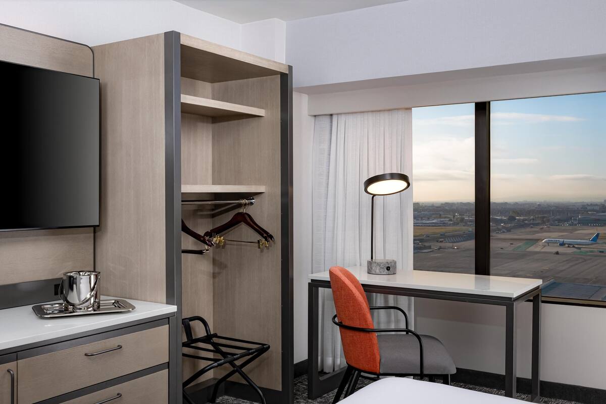 A hotel room with a large window, a desk, a chair, a TV, and a minibar.