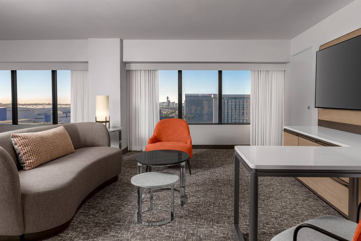 A hotel room with a grey sofa, an orange chair, a small round table, a desk, and a large window offering a city view.