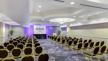 A conference room with rows of chairs, a stage with a Hilton logo, and a chandelier.