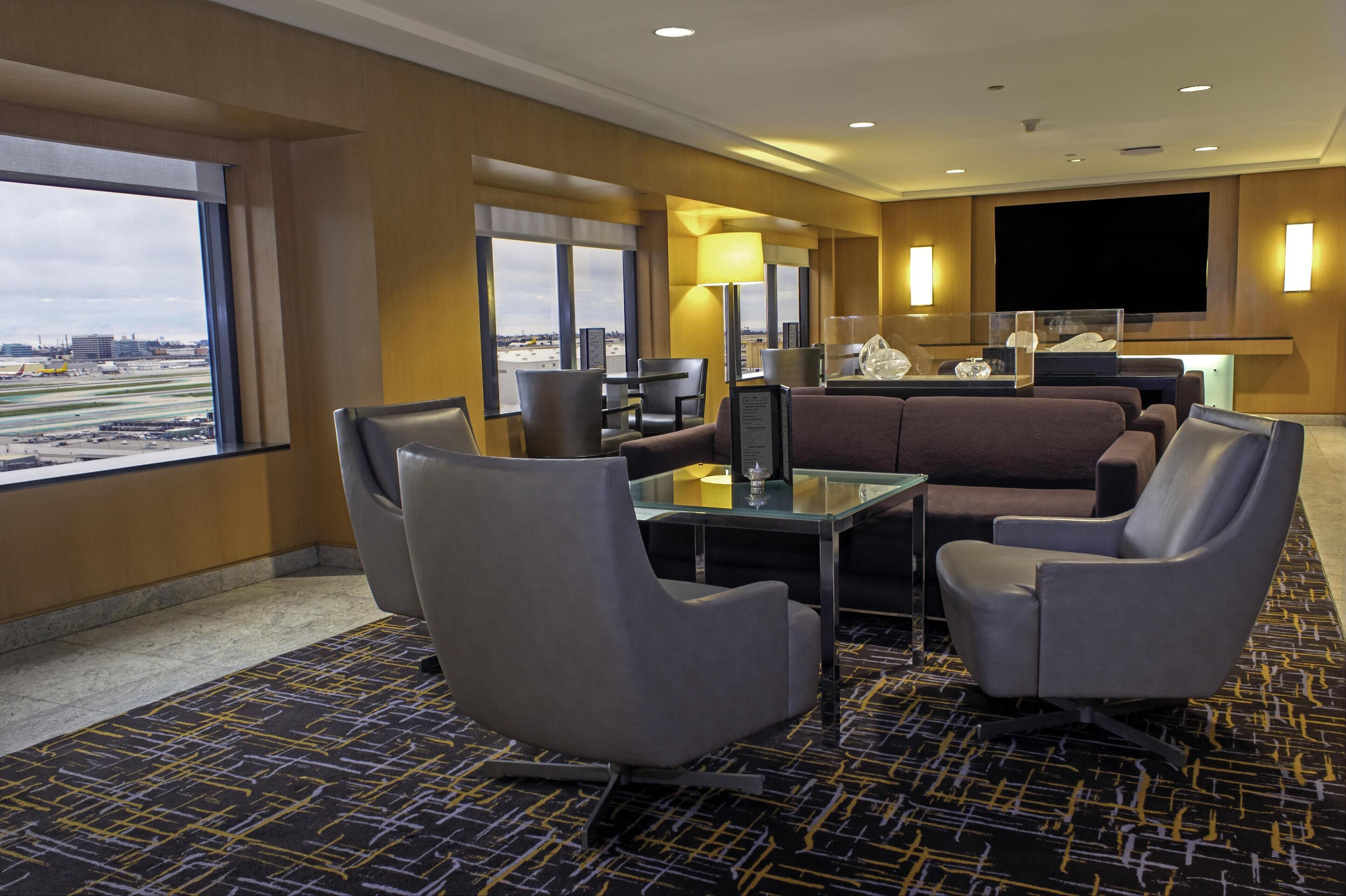 Quarto de hotel com uma grande janela, um sofá, duas poltronas, uma mesa pequena e uma televisão de tela plana fixada na parede.