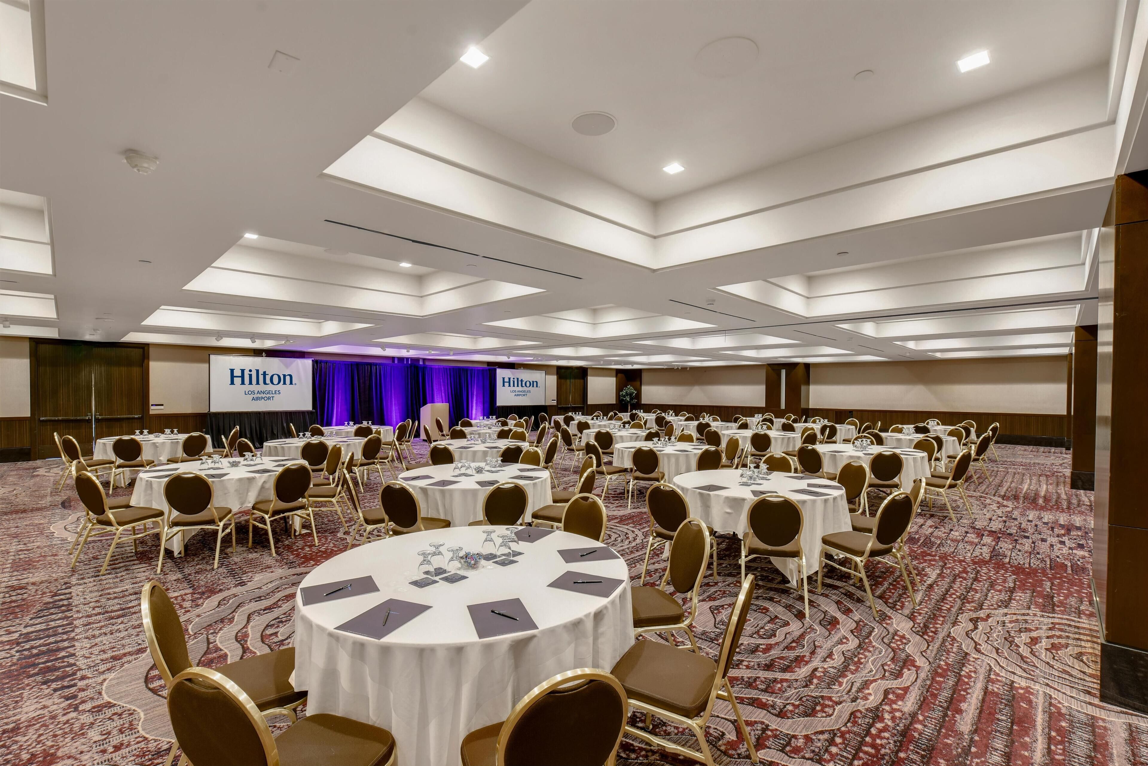 Une grande salle de conférence dotée de plusieurs tables rondes et de chaises, d’un sol recouvert de moquette et d’une scène ornée d’une bannière Hilton.