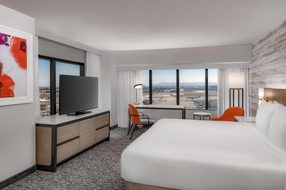 A hotel room with a large bed, a desk with a TV, a chair, and a view of the city.