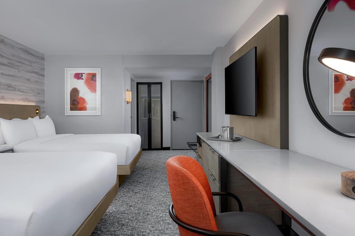 A hotel room with two beds, a desk, a chair, a TV, and artwork on the walls.