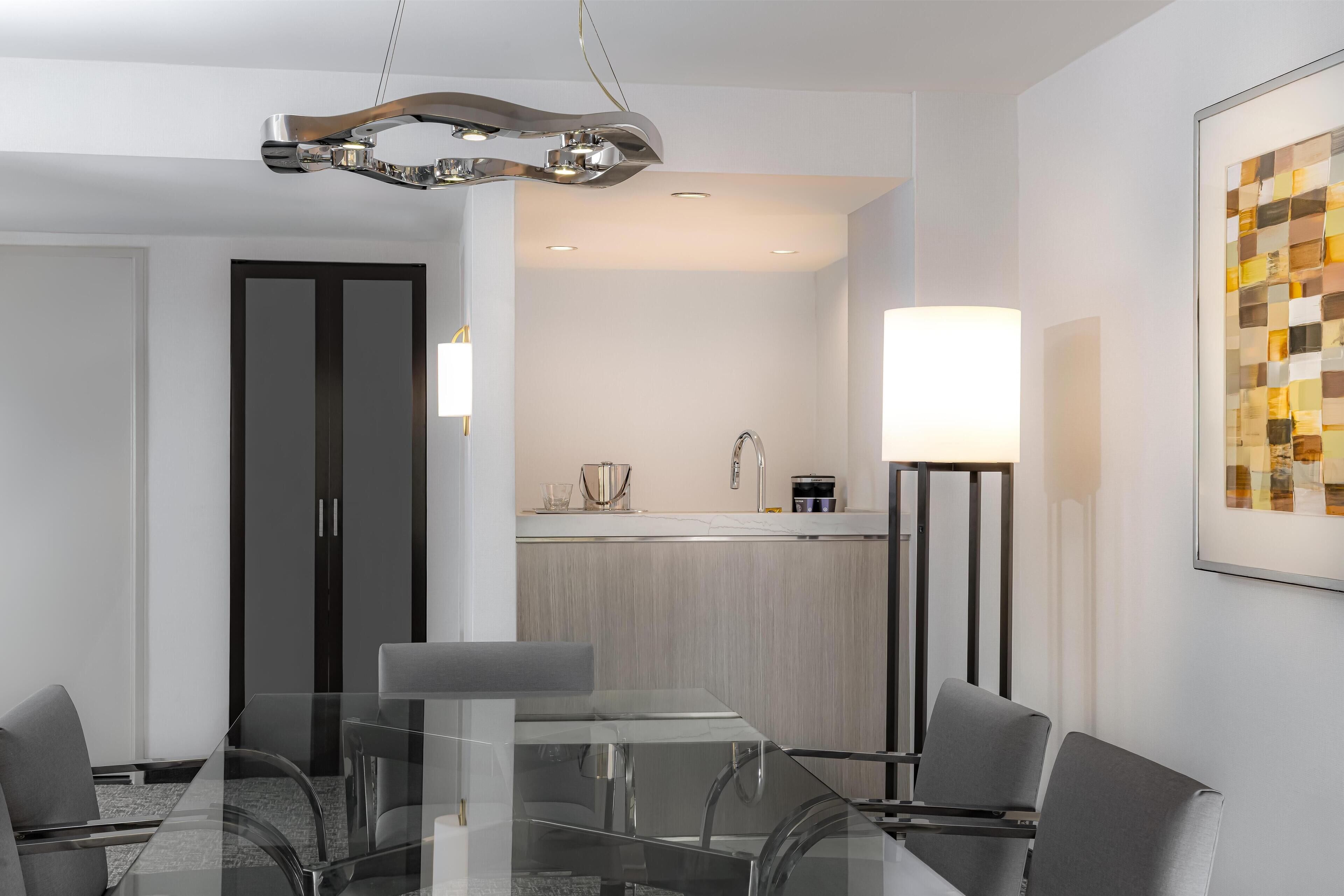 Quarto de hotel moderno com uma mesa de jantar de vidro, cadeiras cinzas, cozinha com pia e um abajur.