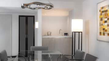 Quarto de hotel moderno com uma mesa de jantar de vidro, cadeiras cinzas, cozinha com pia e um abajur.