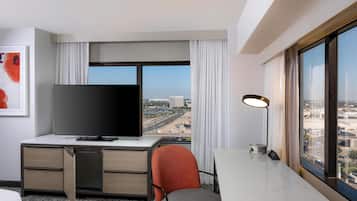 Habitación de hotel con televisor grande, escritorio, silla y vista a la ciudad a través de un ventanal amplio.