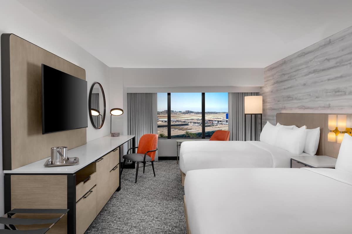 A hotel room with a large TV, two beds, a desk with a chair, and a view of the city.