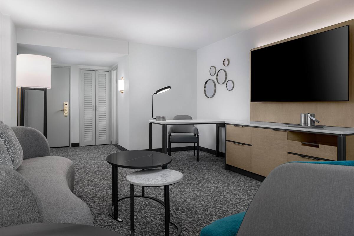 A modern hotel room with a sofa, a small round table, a desk with a chair, a flat-screen TV, and a wall-mounted mirror.