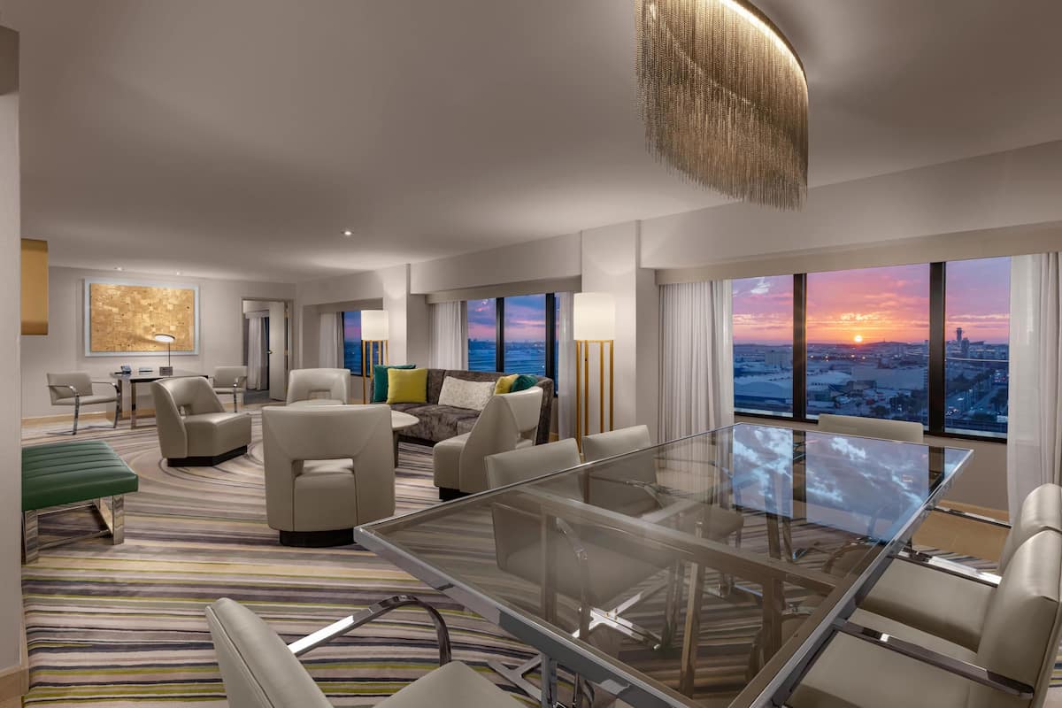 A modern hotel room with a large glass table, comfortable seating, a city view, and a large artwork on the wall.