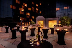 Una terraza al aire libre con mesas dispuestas para comer, rodeada por un edificio moderno con ventanas amplias y una entrada bien iluminada.